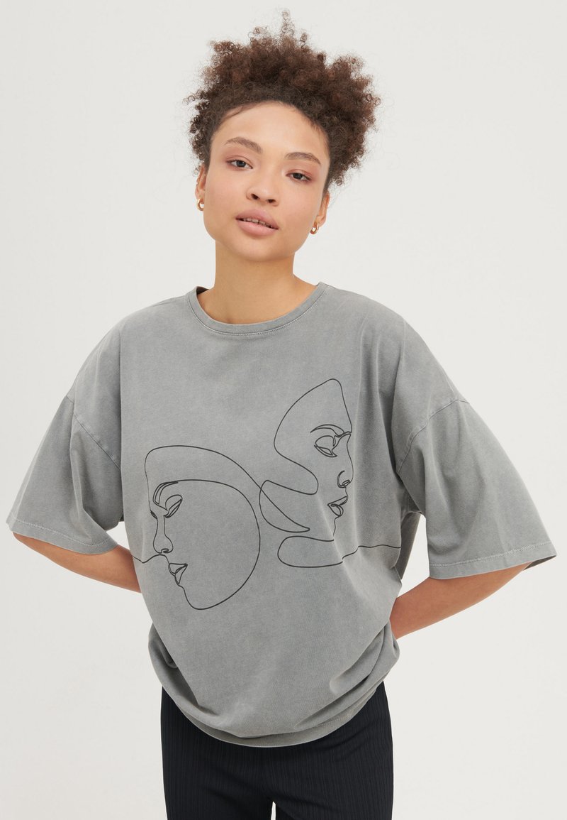 Ung kvinna med lockigt hår bär en oversized grå t-shirt med minimalistisk svart linjekonst föreställande två ansikten, kombinerad med mörka byxor.