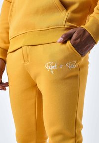 Sudadera amarilla y pantalones deportivos hechos de una mezcla suave de algodón. Presentan puños acanalados y una marca bordada en los pantalones.