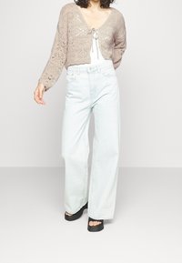 Cárdigan de ganchillo beige con escote en V y cierre con lazo, combinado con jeans de pierna ancha color azul claro y sandalias de plataforma negras.