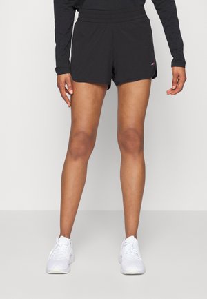 Personne portant un short de sport noir, un haut à manches longues noir et des baskets blanches, se tenant devant un fond clair uni.