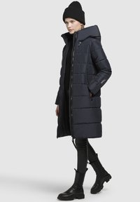 Navy puffer coat with a hood, zip front, and side pockets. Features a quilted design and is worn over black leggings and knee-high boots.