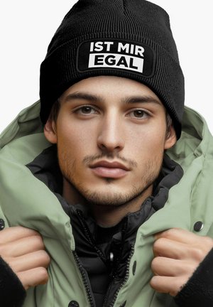 Black knitted beanie with white text patch. Model wears a padded green jacket, visible black inner lining, and grasping jacket fabric.
