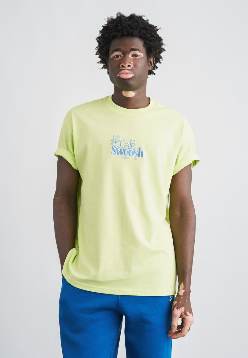 Svetlozelené bavlnené tričko s vyhrnutými rukávmi a logom „Café Swoosh“ v modrej farbe. Model nosí modré tepláky a stojí na neutrálom pozadí.