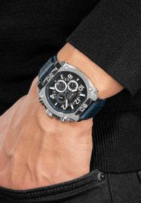 Kraftig silverfärgad klocka med svart urtavla, silvermarkeringar och sub-ur, med ett blått strukturerat läderarmband.