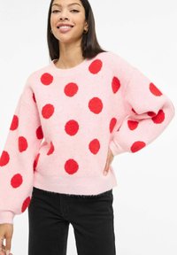 Roosa kampsun suurte punaste karvaste täppidega, ümmarguse kaelusega ja pallikujuliste varrukatega, kombineeritud mustade teksadega. Pehme tekstuur ja lõdvestunud istuvus.