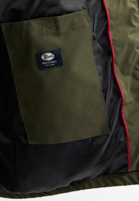Olivegrüner Stoffbeutel mit einem quadratischen Gittermuster, ausgestattet mit einem blauen Markenlabel und schwarzem Satinfutter mit roten Akzenten.
