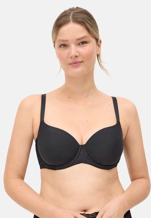 Mujer de piel clara y cabello rubio lleva un sujetador negro ajustado con tirantes regulables sobre un fondo liso.