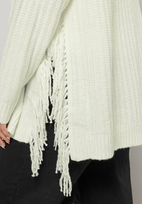 Długi, biały sweter z dzianiny o teksturowanym wzorze, z chwostami na krawędzi. Nosi się go na czarnych spodniach.