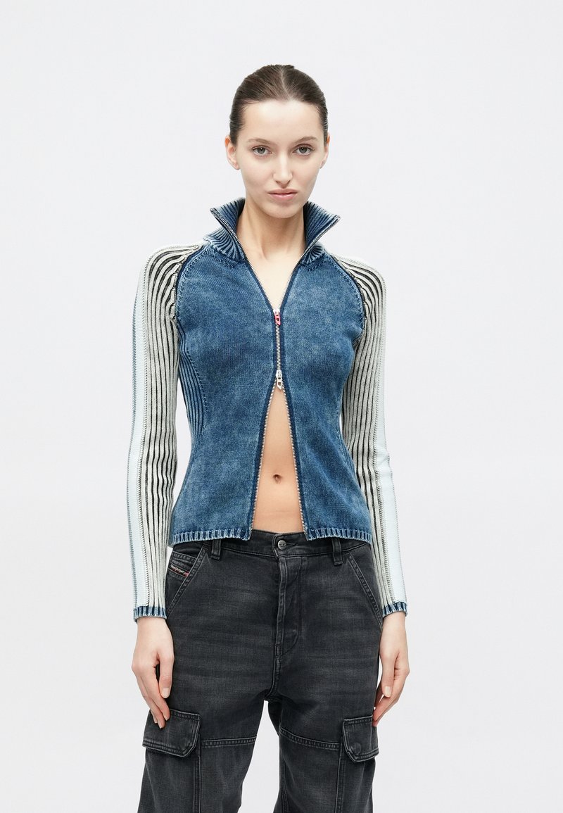 Kvinna som bär en blå tröja med dragkedja framtill och randiga ärmar samt svarta cargobyxor, stående framför en enkel ljus bakgrund.