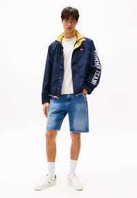 Mørkeblå vindjakke med gul fôr og "TOMMY JEANS"-logo på ermene, sammen med lyseblå denimshorts og hvite sneakers.