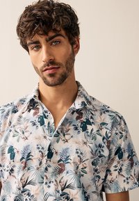 Camicia a maniche corte in tessuto leggero con una combinazione di tonalità verdi, blu e terrose, decorata con un motivo di foglie e fiori tropicali.
