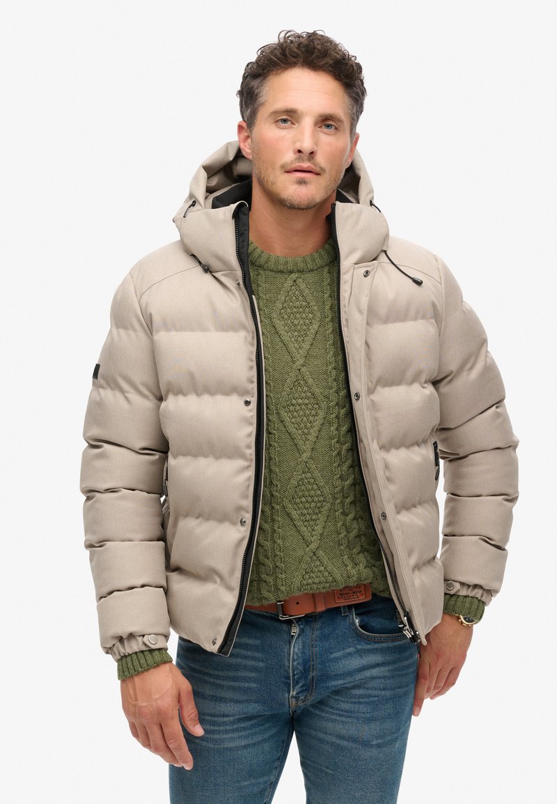 Beige gewatteerde jas met een capuchon, ritssluiting en een ribbeltruien in het groen eronder. Heeft een geborduurd textuur en aansluitende mouwen.