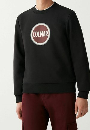 Homme portant un sweat-shirt noir avec un logo circulaire COLMAR bordeaux et blanc sur la poitrine, associé à un pantalon rouge foncé.