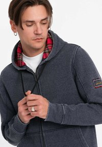 Felpa con zip grigia scura, con un colletto interno a tartan rosso, cordino e tessuto testurizzato. Presenta una toppa sulla manica sinistra.