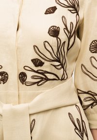 Μπεζ λινό φόρεμα με καφέ ανθιστή κεντητική και φαρδιά υφασμάτινη ζώνη δεμένη στη μέση, διαθέτει μακριά μανίκια και διακριτική ραφή.