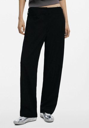 Black wide-leg pants made of soft fabric, featuring a simple design, with pockets and a relaxed fit, paired with silver footwear.