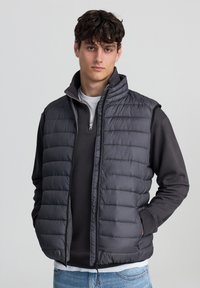 Szary kamizelka puchowa z wysokim kołnierzem, z zamkiem błyskawicznym i poziomymi przeszyciami; noszona na ciemnej bluzie z długim rękawem i białym t-shircie.