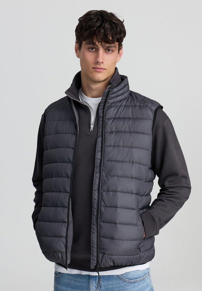 Szary kamizelka puchowa z wysokim kołnierzem, z zamkiem błyskawicznym i poziomymi przeszyciami; noszona na ciemnej bluzie z długim rękawem i białym t-shircie.