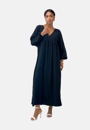 Black long-sleeve dress in textured fabric with a v-neckline and gathered waist. Features a flowy silhouette and is paired with white heeled sandals.
