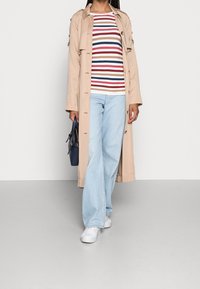 Trench-coat beige sur une chemise à manches longues rayée rouge, bleu et blanc ; jean large bleu clair ; baskets blanches ; sac à main bleu marine.