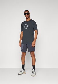 Tumši pelēks sporta t-krekls ar baltu logo, kombinēts ar tumši zilām biksēm ar kabatu. Modelis valkā saulesbrilles un gaiši pelēkus skriešanas apavus.