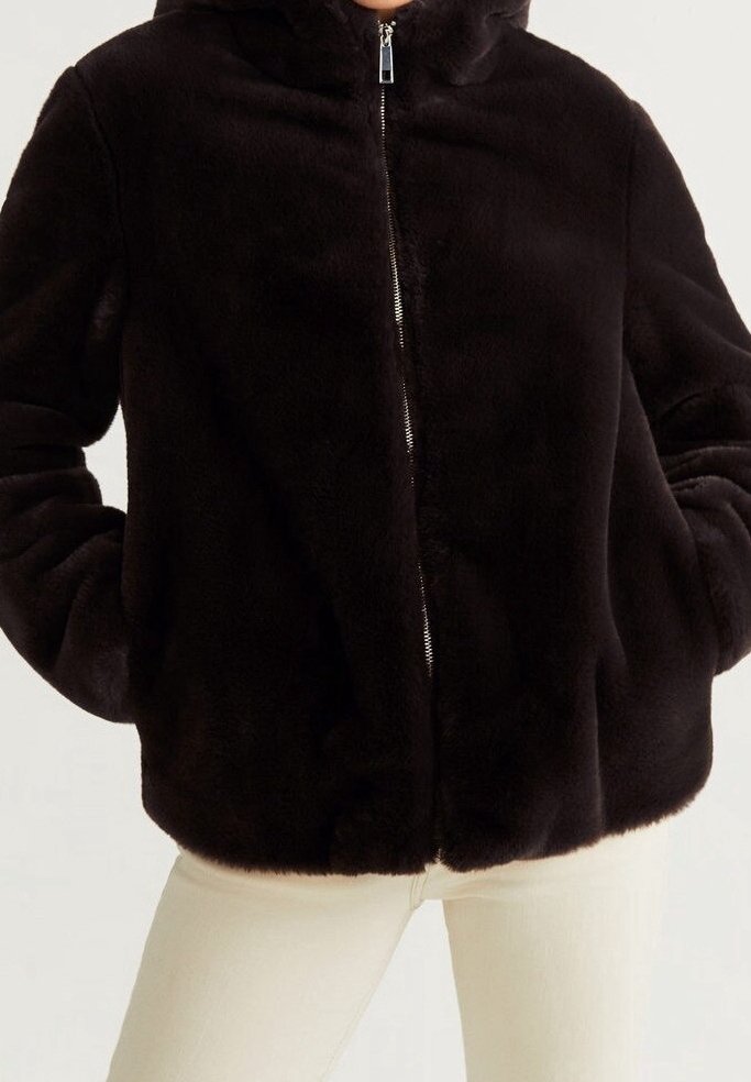 Veste noire en fausse fourrure à zip portée sur un pantalon clair, mains dans les poches de la veste.