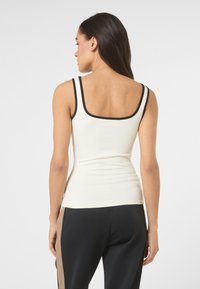 Mujer de pie de espaldas, con una blusa sin mangas blanca con ribete negro y pantalones negros con franjas beige en los laterales.