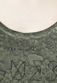 Camiseta verde olivo con cuello redondo; presenta un tejido texturizado y el texto "El mundo de" sobre un patrón de logo con diamantes.