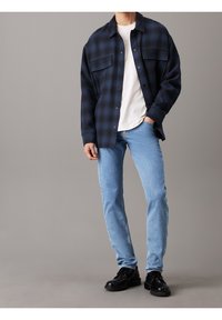 Modrá kostkovaná oversized košile, pod ní bílá tričko, světlé modré džíny a černé boty. Košile má dvě kapsy na prsou a knoflíky.