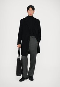 Uomo in soprabito nero, dolcevita nero, pantaloni grigi, scarpe nere, che tiene una borsa tote nera, in piedi contro uno sfondo bianco.