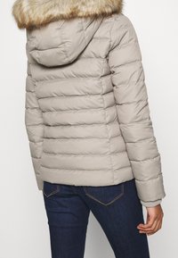 Pufferjakke i lys beige med horisontalt kviltet mønster. Har en faux fur hættekanter og ribbede manchetter. Sammen med mørkeblå jeans.