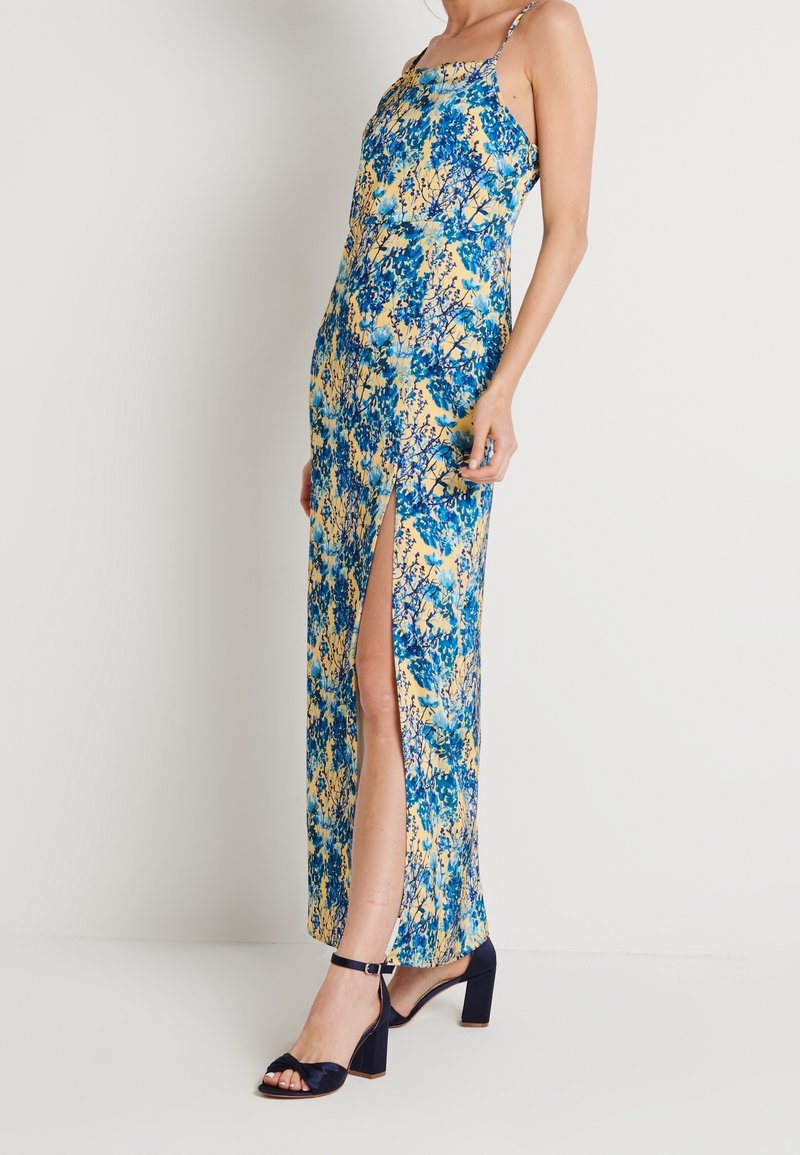 Blumenmaxikleid mit blauen und gelben Mustern, dünnen Trägern, seitlichen Schlitzen und lockerer Passform. Kombiniert mit schwarzen Sandalen mit Blockabsatz.