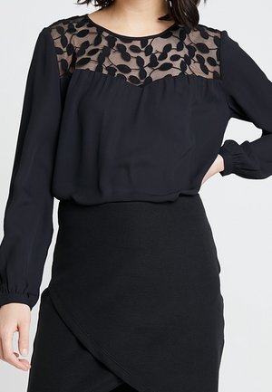 Kobieta ubrana w czarną bluzkę z długim rękawem, z prześwitującym karczkiem w liściasty wzór oraz dopasowaną czarną spódnicę z asymetrycznym dołem.