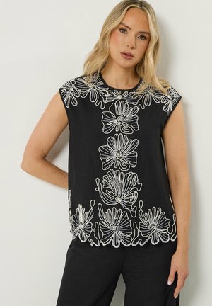 Blondynka w czarnym topie bez rękawów ozdobionym białym kwiatowym haftem wzdłuż dekoltu, środka i brzegu, w zestawieniu z czarnymi spodniami.