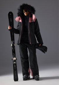 Skiklær som inkluderer en sort og rosa isolert jakke med pelskant, svarte skibukser, skibriller og svarte hansker, som holder svarte ski.