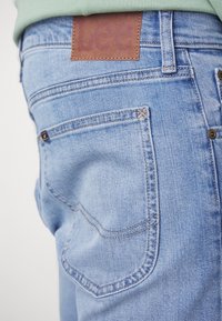 Dos de jean denim bleu clair avec une étiquette de marque en cuir marron et des coutures visibles, porté avec un haut vert clair.