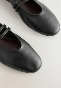 Musta nahka ballet-kengät, joissa on pyöreä kärki ja kaksi ohutta hihnaa päältä. Pinta on sileä ja siinä on hienovarainen kiilto.