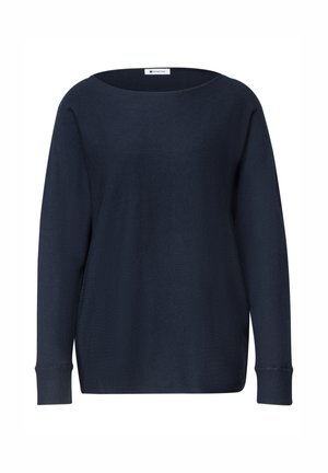 Donna con capelli alla lunghezza delle spalle che sorride, indossa una maglia a maniche lunghe blu navy e jeans azzurri, in piedi contro uno sfondo semplice.