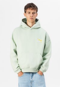 Lys grønn hettegenser med frontlomme, avslappet passform, brodert "On Vacation" design i gult, og ribbestrikkede mansjetter og nederkant.