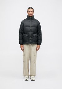 Mann med flettehår iført en svart puffjakke, beige cargo-bukser og hvite sneakers som står mot en ensfarget hvit bakgrunn.