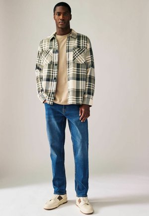 Geruite flanellen shirt in crème, groen en marineblauw over een beige t-shirt; blauwe spijkerbroek en beige sneakers met bruine accenten.