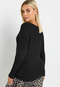 Vaalea nainen, joka on pukeutunut mustaan ribbipintaiseen pitkähihaiseen neuleeseen ja leopardikuvioisiin housuihin, katsoo olkansa yli valkoista taustaa vasten.