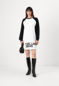 Vestido branco com mangas raglan negras, apresentando um atrevido logo preto "KARL LAGERFELD JEANS" na barra. Combinado com botas pretas até ao joelho.