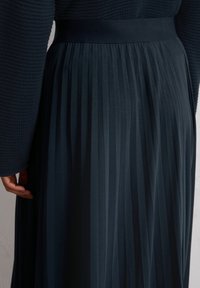 Un'indicazione parziale di una persona che indossa un maglione scuro dalla texture interessante e una gonna scura fluente e plissettata, evidenziando il drappeggio e il movimento del tessuto.