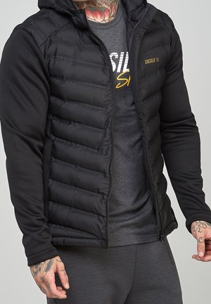 Mężczyzna w czarnej, pikowanej kurtce z kapturem, ciemnoszarej koszulce Siksilk oraz ciemnoszarych spodniach, z widocznymi tatuażami na dłoniach i szyi.