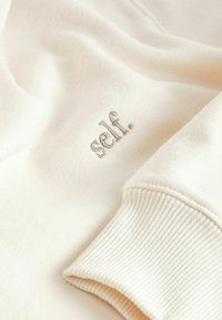 Cream-colored sweatshirt with ribbed cuffs and the embroidered word "self." in a subtle beige. Soft texture and relaxed fit.