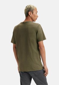 Mand med kort blond hår iført en olivengrøn t-shirt og mørkegrå jeans, vendt sidelæns mod en ensfarvet baggrund.