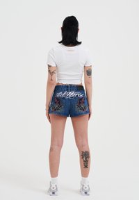 Denim shorts met kleurrijke bloemborduursels en de tekst "Cold Stare" op de achterkant, gecombineerd met een witte cropped t-shirt en witte sportsokken.