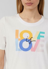 T-shirt en coton blanc avec un graphique multicolore portant le texte "LOVE is LOVE" dans différentes couleurs et polices, avec une texture douce et lisse.