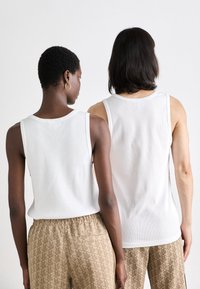 Hvite ribbede tanktops på to modeller, med brede stropper og rund hals, sammen med mønstrede beige bukser.